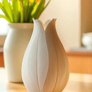 May include: A white, tulip-shaped vase with textured, petal-like details. The vase is on a wooden surface, with a blurred background featuring a white vase holding pink and yellow flowers. The vase is a decorative item.