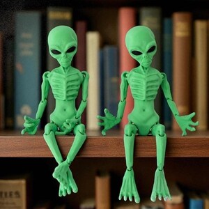 May include: Two articulated green alien figures with large black eyes sit on a wooden shelf. The figures have detailed rib cages and are posed with their arms and legs extended. The background is a blurred collection of books.