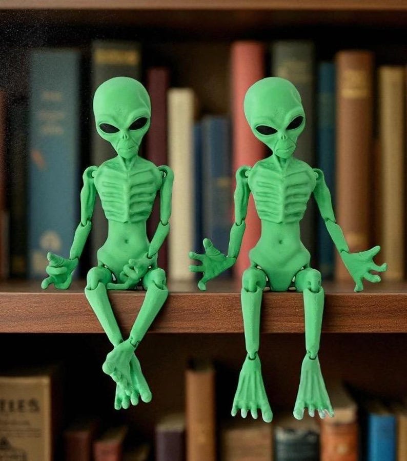 May include: Two articulated green alien figures with large black eyes sit on a wooden shelf. The figures have detailed rib cages and are posed with their arms and legs extended. The background is a blurred collection of books.