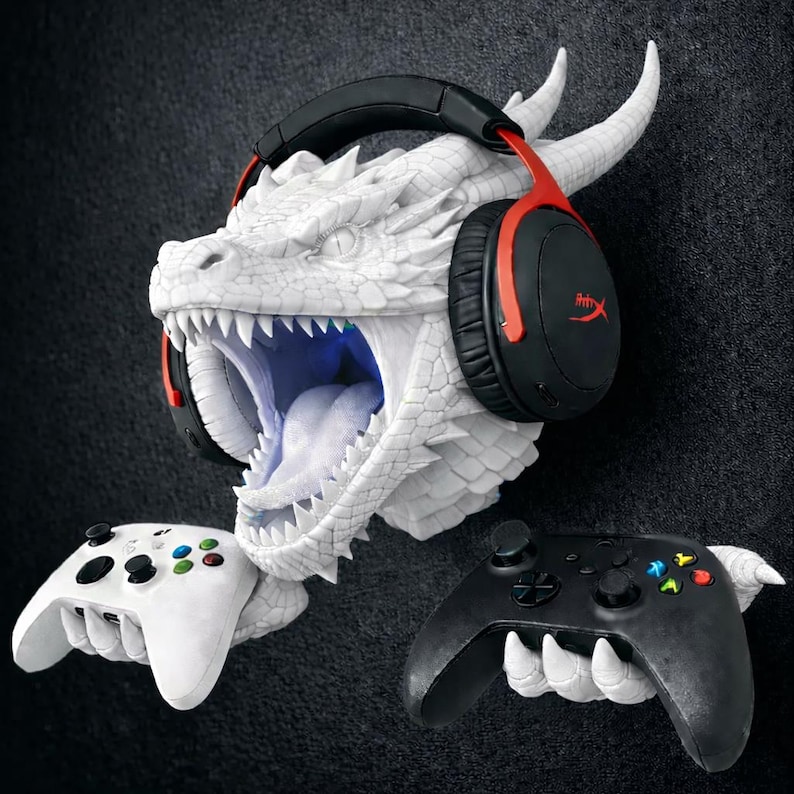 May include: A white dragon head wall mount with a headset and two game controllers. The dragon's mouth is open, revealing teeth and a blue interior. The headset is black and red. One controller is white, the other is black.