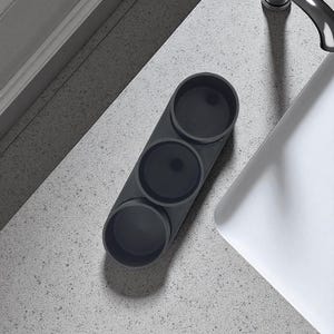 May include: A dark grey, three-section organiser for small items. The cylindrical compartments are arranged linearly. The organiser sits on a speckled, light grey surface near a white sink.