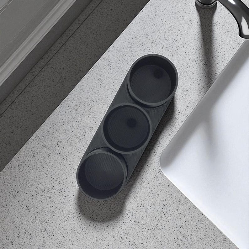 May include: A dark grey, three-section organiser for small items. The cylindrical compartments are arranged linearly. The organiser sits on a speckled, light grey surface near a white sink.