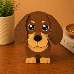 K&ouml;nnte beinhalten: Eine braune und beige Dackel-Hundefigur mit gro&szlig;en, ausdrucksstarken Augen und Schlappohren. Der Hund hat eine schwarze Nase und steht aufrecht. Die Figur steht auf einer Holzoberfl&auml;che, mit einer Pflanze und einem Buch im Hintergrund.