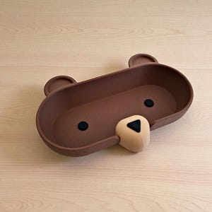 Op de afbeelding: Een bruine, beer-vormige tray met twee kleine oren, twee zwarte ogen en een beige neus. De tray is ovaal van vorm en gemaakt van een mat materiaal. De neus van de beer heeft een zwarte driehoek. De tray staat op een licht houten oppervlak.