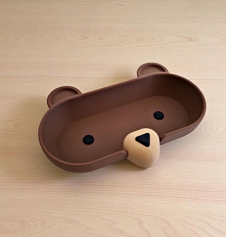 Op de afbeelding: Een bruine, beer-vormige tray met twee kleine oren, twee zwarte ogen en een beige neus. De tray is ovaal van vorm en gemaakt van een mat materiaal. De neus van de beer heeft een zwarte driehoek. De tray staat op een licht houten oppervlak.