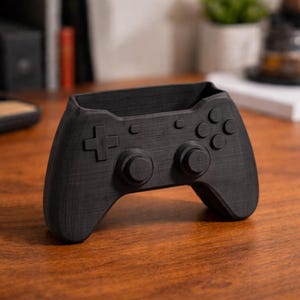 May include: A black pen holder in the shape of a video game controller. The controller has a directional pad, buttons, and analog sticks. The pen holder is made of a dark material and is on a wooden surface.