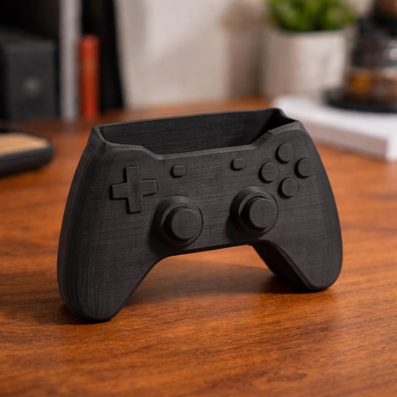 May include: A black pen holder in the shape of a video game controller. The controller has a directional pad, buttons, and analog sticks. The pen holder is made of a dark material and is on a wooden surface.