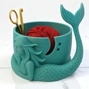 May include: A teal mermaid-themed yarn bowl with a red ball of yarn inside. The bowl has a mermaid tail and a sculpted mermaid figure on the side. A gold crochet hook and scissors are placed inside the bowl.