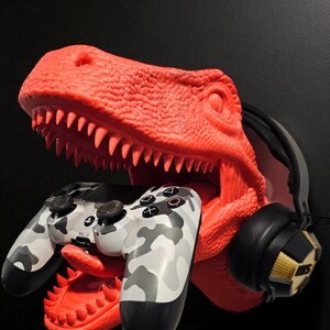 May include: A red dinosaur head sculpture holds a camouflage-patterned game controller in its mouth. A pair of black and gold headphones rests on the side. The sculpture is a unique gaming accessory.