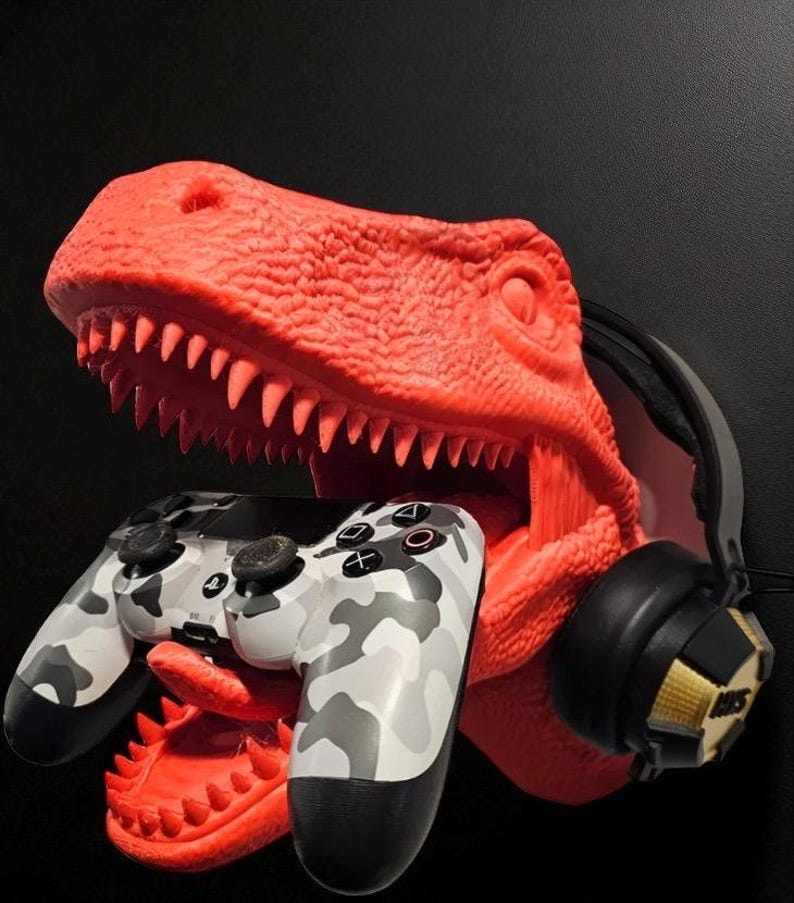 May include: A red dinosaur head sculpture holds a camouflage-patterned game controller in its mouth. A pair of black and gold headphones rests on the side. The sculpture is a unique gaming accessory.