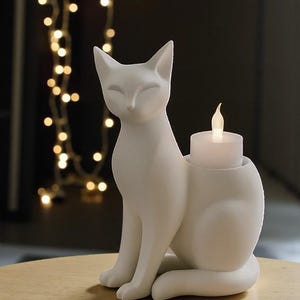 Op de afbeelding: Een witte keramische kattenfiguur met een kandelaar. De kat zit met gesloten ogen. Een witte kaars met een flikkerende vlam staat in de houder. De achtergrond is donker met lichtslingers.