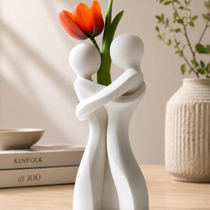 May include: A white ceramic vase in the shape of two embracing figures, holding a vibrant orange tulip with green leaves. The vase is on a wooden surface, with a book and a decorative vase in the background.