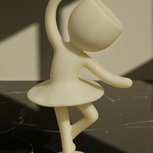 May include: A cream-colored figurine of a ballerina in a graceful pose. The figure has a rounded head, arms outstretched, and a tutu. The figurine is standing on one leg, with the other bent.