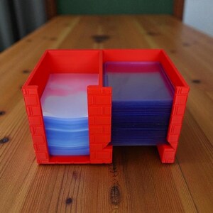 May include: A red plastic card holder with two compartments. One side holds a stack of blue-tinted card sleeves, and the other holds a stack of white and blue card sleeves. The holder has a brick-like design on the sides.