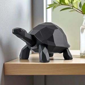 May include: A black geometric turtle sculpture with a faceted design. The decorative figurine is made of a solid material and has a smooth, matte finish. The turtle is displayed on a light-colored wooden shelf.
