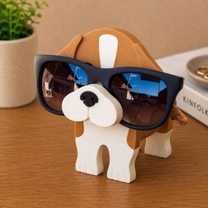 May include: A whimsical dog-shaped sunglasses holder. The holder is primarily white and brown, with a black nose and a pair of dark blue sunglasses. The sunglasses reflect the surrounding environment. The holder is made of a solid material and is designed to hold eyewear.