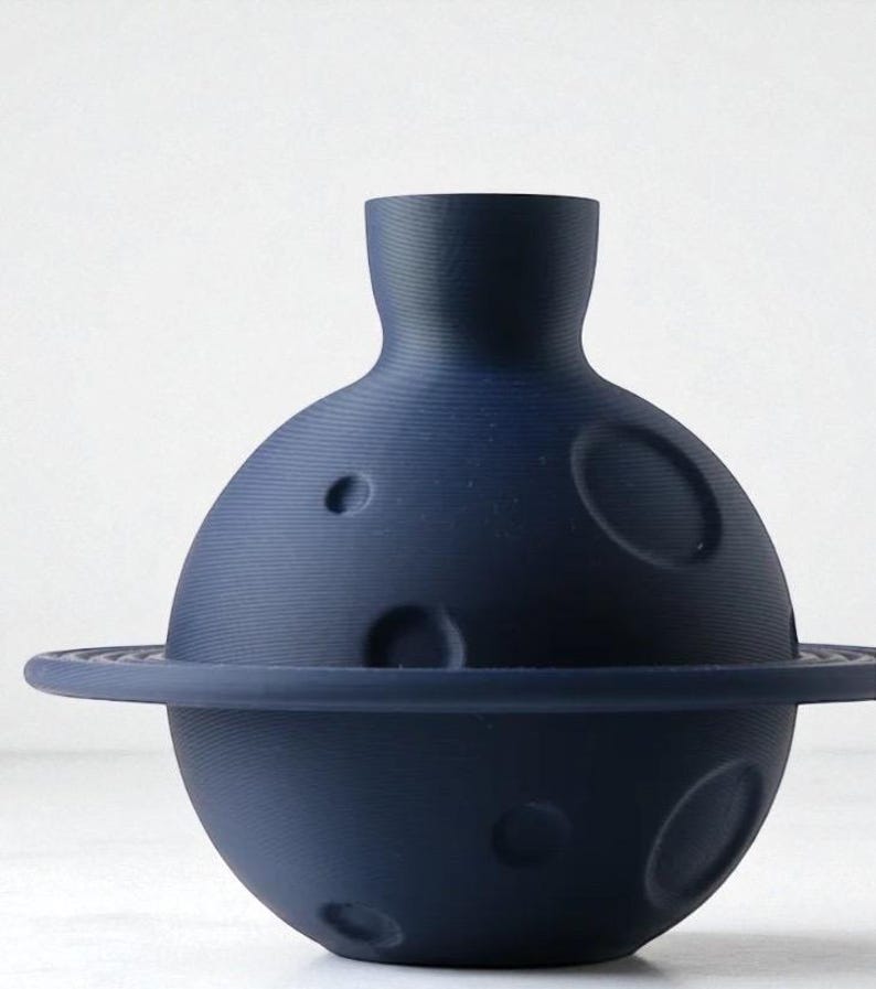 Może przedstawiać: Granatowy ceramiczny wazon w kształcie planety z pierścieniem. Wazon ma okrągły korpus z kilkoma okrągłymi wklęsłościami i cylindryczną szyjką. Pierścień otacza środkową część planety.