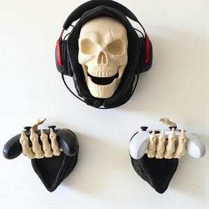 Puede incluir: Una calavera crema con una sudadera con capucha negra y auriculares para juegos con detalles en rojo. Debajo, dos soportes de tela negra presentan manos esqueléticas agarrando mandos de videojuegos, uno negro y otro blanco.