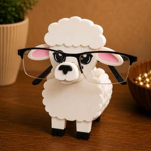 May include: A white sheep-shaped eyeglasses holder with black-rimmed glasses. The sheep has pink inner ears and black hooves. The holder is made of a solid material and sits on a wooden surface. A decorative item for a desk or nightstand.
