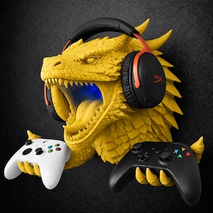 May include: A yellow dragon head wall mount featuring a textured scale pattern and an open mouth with a blue light. The dragon head wears black and red over-ear headphones and uses its clawed hands to hold a white game controller and a black game controller.