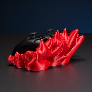 May include: A vibrant red, flame-shaped gaming controller stand. The stand is in front of two black game controllers. The stand is made of a shiny, smooth material and is on a dark surface.