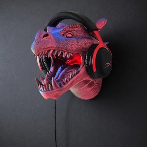 May include: A red and blue dragon head wall mount with a pair of black and red headphones. The dragon's mouth is open, revealing sharp teeth and a red glowing interior. The headphones have the HyperX logo.