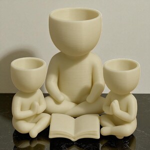 May include: Three cream-colored figurines in a meditative pose. The central figure is larger, with two smaller figures flanking it. An open book rests in front. The figures have open-topped heads, suggesting they are planters or containers.