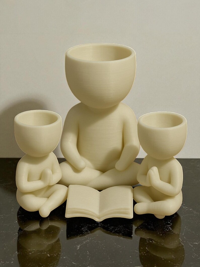 May include: Three cream-colored figurines in a meditative pose. The central figure is larger, with two smaller figures flanking it. An open book rests in front. The figures have open-topped heads, suggesting they are planters or containers.