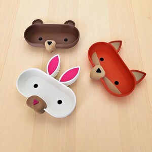 May include: Three animal-shaped trays: a brown bear, a white rabbit with pink ears, and an orange fox. Each tray has a long oval shape with the animal's face at one end. The bear has a tan nose, the rabbit has a brown nose, and the fox has a tan nose.