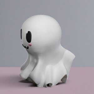 May include: A white ghost figurine with a rounded head and a draped body. The ghost has black oval eyes, a small black mouth, and pink blush marks on its cheeks. The figurine is set against a gray and pink background.