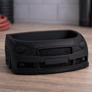 May include: A matte black pen holder shaped like the front of a car. The holder features headlights, a grille, and a bumper, and is designed to sit on a desk or table. The product is made of a solid material.