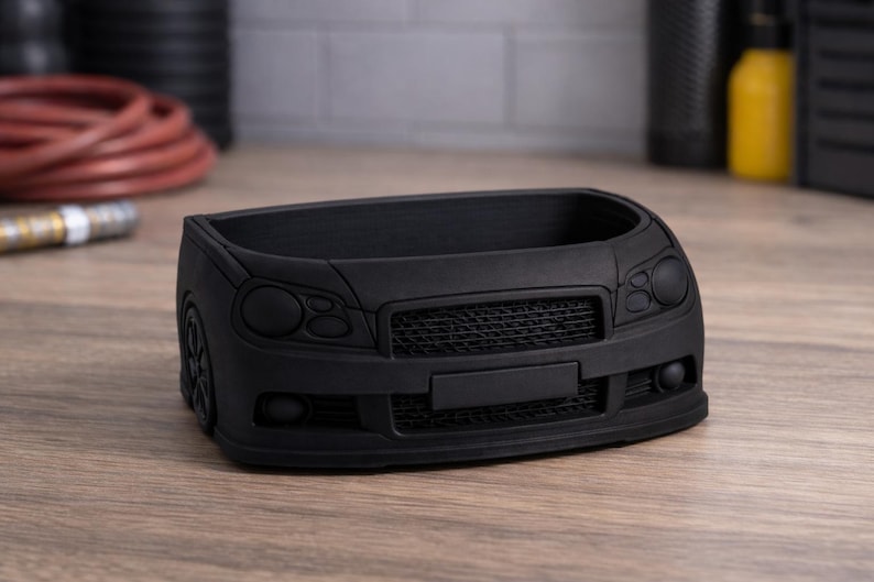 May include: A matte black pen holder shaped like the front of a car. The holder features headlights, a grille, and a bumper, and is designed to sit on a desk or table. The product is made of a solid material.