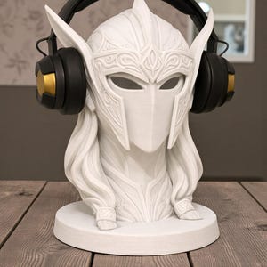 May include: A white headphone stand in the form of a warrior's head, featuring a detailed helmet and flowing hair. Black and gold headphones are placed on the head. The stand is on a round base, ideal for showcasing headphones.