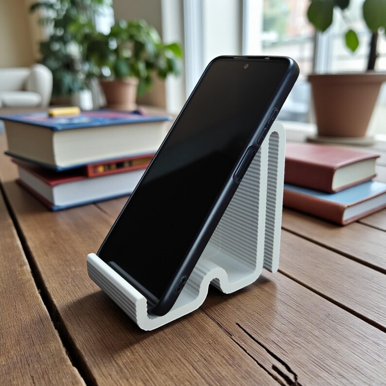 Op de afbeelding: Een witte, geribbelde telefoonstandaard met een zwarte smartphone. De standaard staat op een houten tafel, met boeken en planten op de achtergrond. De standaard is ontworpen om een telefoon in een hoek te houden.