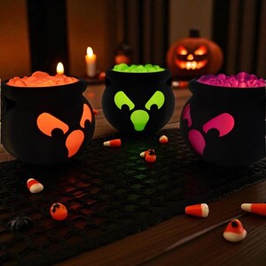 May include: Three black cauldron-shaped containers with glowing faces and filled with colorful candies. The cauldrons have orange, green, and pink faces. Candy corn is scattered around the cauldrons. A lit jack-o'-lantern is in the background, creating a Halloween theme.