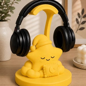 May include: A yellow star-shaped headphone stand with a sleeping cap and a crescent moon design. Black headphones rest on the stand. The stand is on a round base. The overall design is whimsical and decorative.