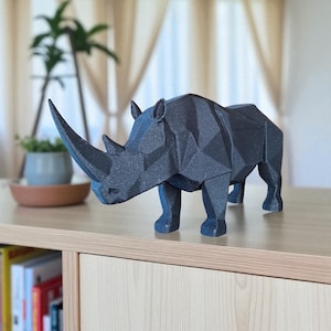 May include: A dark gray geometric rhino sculpture with a long horn, standing on a light wood surface. The rhino is made of angular shapes, giving it a modern, artistic look. The sculpture is a decorative item.