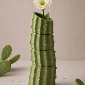 May include: A green cactus-shaped vase with a white flower and green leaves. The vase is made of stacked sections, with a textured surface and small, pointed details. The flower is in full bloom, with delicate petals.