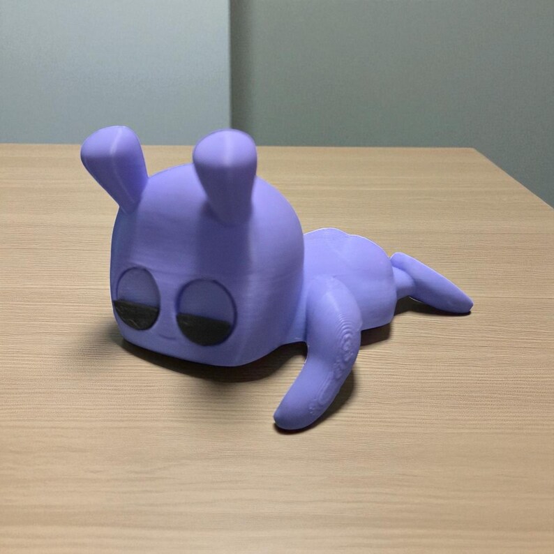Pu&ograve; includere: Figurina lavanda stampata in 3D di una creatura stilizzata. La figura ha una testa arrotondata con due orecchie dritte, grandi occhi ovali e un corpo che si assottiglia in una coda. La creatura &egrave; sdraiata sulla pancia con le braccia e le gambe distese.