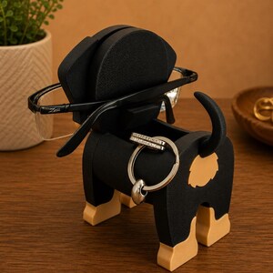 May include: A black and tan dog-shaped organizer holding eyeglasses and a ring. The organizer has a compartment for small items and a slot for glasses. The dog's tail is curved upwards, and the legs are tan.
