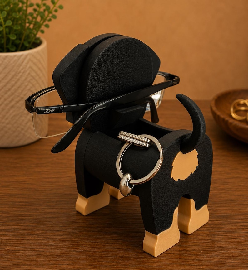 May include: A black and tan dog-shaped organizer holding eyeglasses and a ring. The organizer has a compartment for small items and a slot for glasses. The dog's tail is curved upwards, and the legs are tan.