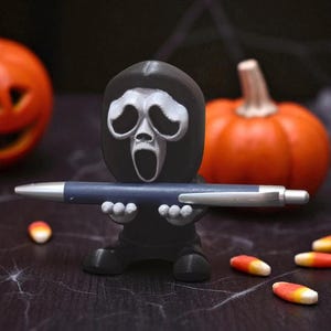Puede incluir: Una figura negra de Ghostface sosteniendo un bol&iacute;grafo azul. Dos calabazas naranjas en el fondo. Caramelos de ma&iacute;z esparcidos. Un portal&aacute;pices con tem&aacute;tica de Halloween.