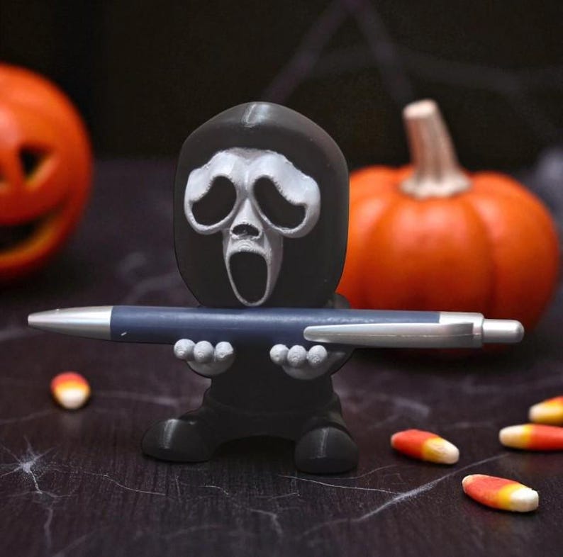 Puede incluir: Una figura negra de Ghostface sosteniendo un bol&iacute;grafo azul. Dos calabazas naranjas en el fondo. Caramelos de ma&iacute;z esparcidos. Un portal&aacute;pices con tem&aacute;tica de Halloween.