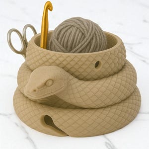 May include: A tan-colored snake-shaped yarn bowl with a ball of yarn, a gold crochet hook, and scissors. The snake's body coils around the bowl, with a detailed scale pattern. The bowl holds the yarn and tools for knitting or crocheting.