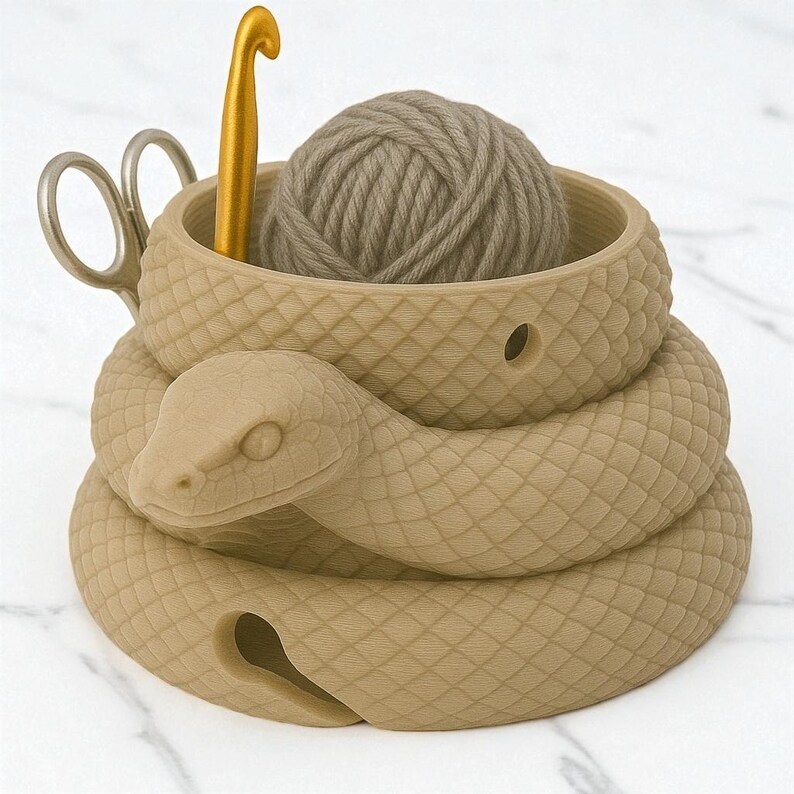 May include: A tan-colored snake-shaped yarn bowl with a ball of yarn, a gold crochet hook, and scissors. The snake's body coils around the bowl, with a detailed scale pattern. The bowl holds the yarn and tools for knitting or crocheting.
