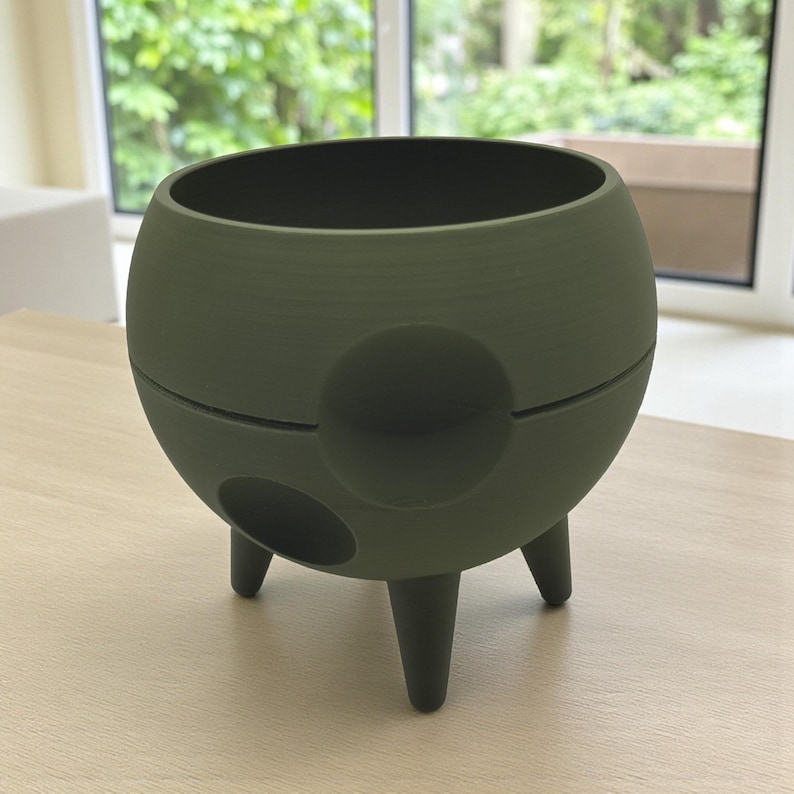 Puede incluir: Una maceta esf&eacute;rica verde oliva oscuro con tres patas cortas y dos aberturas circulares. La maceta tiene una superficie texturizada y una amplia abertura en la parte superior. El dise&ntilde;o es moderno y minimalista.