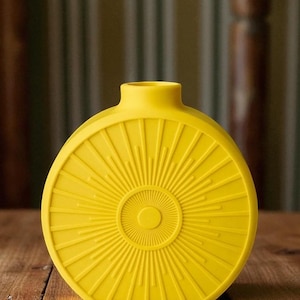 May include: A bright yellow, round vase with a sunburst design. The vase has a narrow neck and a flat base. The sunburst pattern radiates from a central circle, creating a striking visual effect. The vase is made of a solid material.