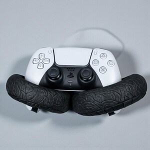 May include: A white and black PlayStation 5 controller is cradled by two black, textured, tire-shaped objects. The controller features a symmetrical design with various button shapes and a central touchpad.