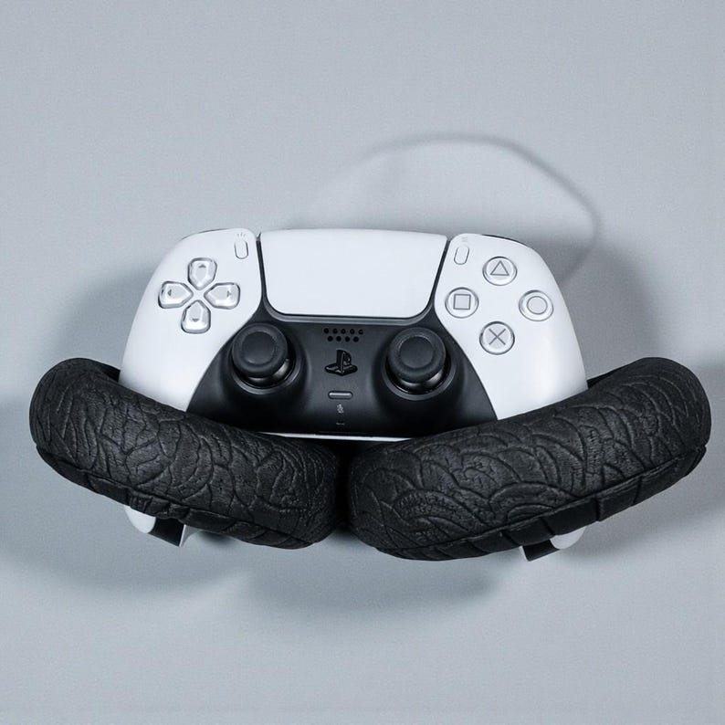 May include: A white and black PlayStation 5 controller is cradled by two black, textured, tire-shaped objects. The controller features a symmetrical design with various button shapes and a central touchpad.
