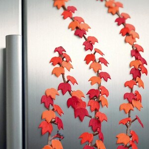 May include: Decorative fall-themed leaf vines in shades of red and orange. The artificial leaves are attached to brown stems, creating a realistic autumn aesthetic. The vines are arranged vertically against a metallic background.
